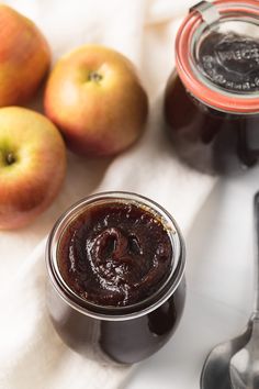 two jars of jam sit next to some apples and spoons on a white cloth