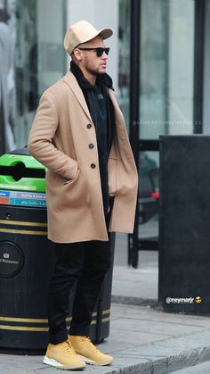 a man in a tan coat and hat standing next to a trash can