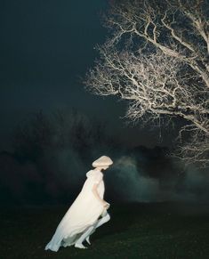 a woman standing in front of a tree at night wearing a white dress and hat