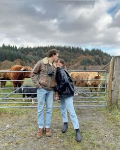 two people standing in front of a fence with cows behind them on a cloudy day