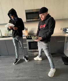 two young men standing in a kitchen looking at something on the counter top and one is wearing a black hoodie