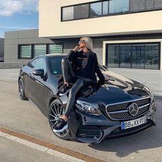 a woman sitting on the hood of a black sports car in front of a building