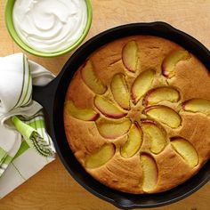 a cake in a pan with apples on top and whipped cream next to it, sitting on a wooden table