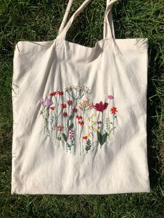 a white tote bag with flowers painted on it sitting in the grass next to a pair of scissors