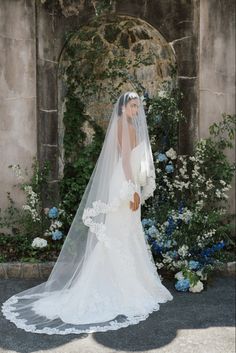 a woman in a wedding dress and veil standing next to blue flowers with an archway behind her