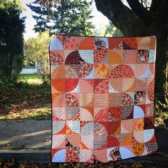 an orange and white quilt sitting on the ground next to a tree with lots of leaves