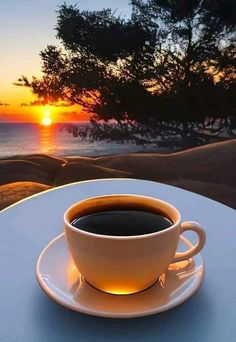 a cup of coffee sitting on top of a white table next to the ocean at sunset