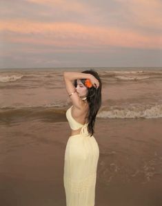 a woman in a yellow dress standing on the beach with her hands behind her head