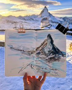 someone is holding up an open book in front of the snow covered mountains and sky