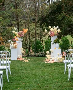 an outdoor ceremony setup with white chairs and floral arrangements on the grass in front of trees