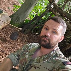 a man with a beard sitting in the woods next to a fire hydrant and trees