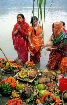 three women in sari are standing by the water with fruits and vegetables on display