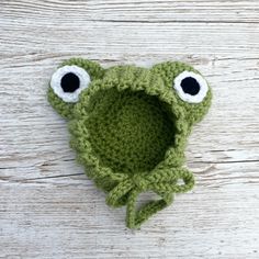 a crocheted frog hat with googly eyes sits on a white wooden surface