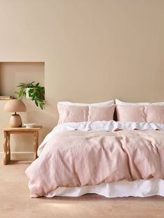 a bed with pink linens and pillows in a neutral colored room, next to a plant