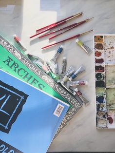 an assortment of art supplies sitting on a table next to some paintbrushes and watercolors