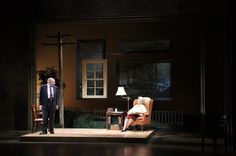 two people are standing on stage in front of a chair and table with a lamp