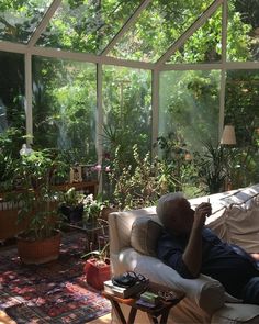 an older man laying on a couch in front of a glass house with lots of plants