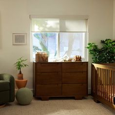 a baby crib in front of a window next to a green chair and potted plant