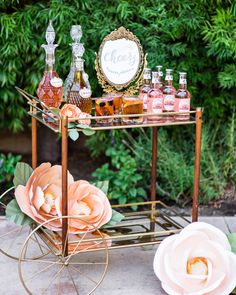 a cart with bottles and flowers on it sitting in front of some bushes next to a pink flower