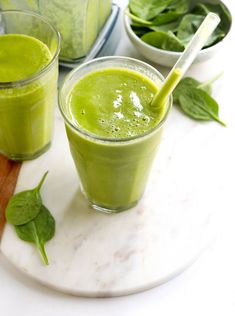 two glasses filled with green smoothie on top of a white plate next to spinach leaves