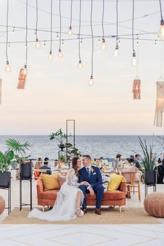 a bride and groom sitting on an orange couch in front of the ocean at sunset