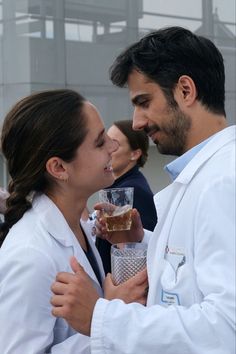 two people in white lab coats are kissing each other and holding glasses with liquid on them