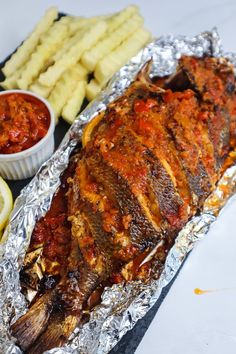 a fish covered in sauce next to french fries and ketchup on tin foil