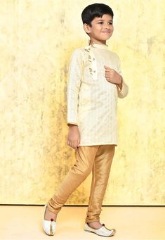 a young boy standing in front of a wall wearing a white shirt and gold pants