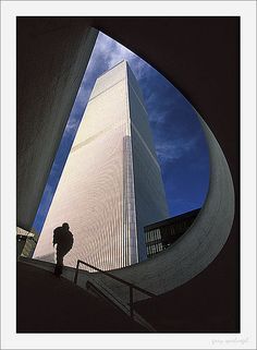 a man standing in front of a tall building with a blue sky behind him and his shadow on the ground