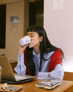 a woman sitting at a table drinking from a cup next to a laptop computer on top of a wooden desk