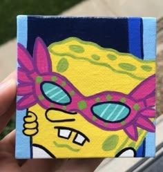 a hand holding up a small square piece of art that looks like spongebob