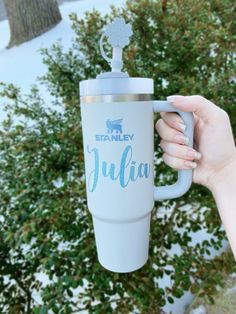 a person holding up a travel mug in front of some bushes and trees with snow on the ground