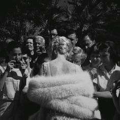 a black and white photo of a woman in a fur coat surrounded by other people