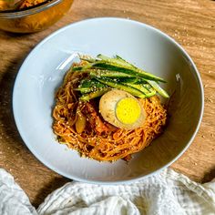 a white bowl filled with noodles and topped with an egg on top, sitting on a wooden table