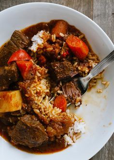 a white bowl filled with meat and rice on top of a wooden table next to a fork