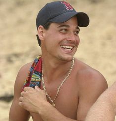 a shirtless man sitting on the beach wearing a baseball cap and scarf around his neck