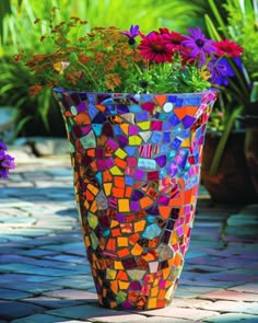 a colorful mosaic vase filled with flowers on top of a stone walkway next to potted plants