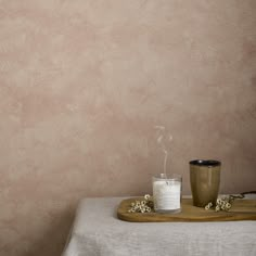 a candle on a table with a glass and some flowers in front of the wall