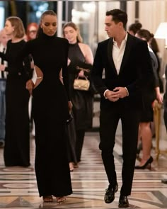 a man and woman dressed in black walking down a hall together, with other people looking on