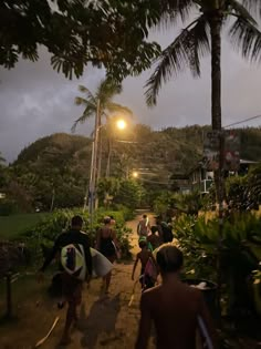 a group of people carrying surfboards down a dirt path at night with palm trees in the background