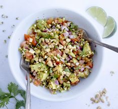 a white bowl filled with chopped vegetables and garnished with cilantro, avocado, and lime