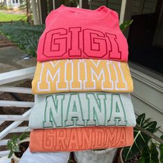 a person holding four different colored shirts in front of a house with the words, gigi, mmmm, nanna and grandma written on them