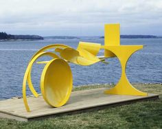 a yellow sculpture sitting on top of a wooden platform next to the ocean and trees