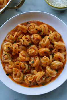 a white bowl filled with pasta and sauce on top of a marble table next to two silver spoons