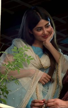 a woman wearing a white sari and holding her hand to her face while standing next to a plant