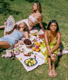 three women laying on a blanket in the grass with fruit and flowers around them,