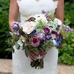 a bride holding a bouquet of purple and white flowers on her wedding day in front of greenery