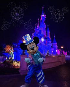 mickey mouse in front of the disneyland castle at night with lights and fireworks behind him