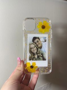 a hand holding a clear case with yellow flowers on it and a photo in the back