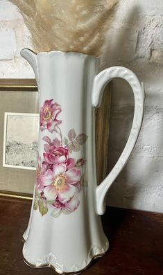 a white vase with pink flowers painted on it sitting on a table next to a framed photo